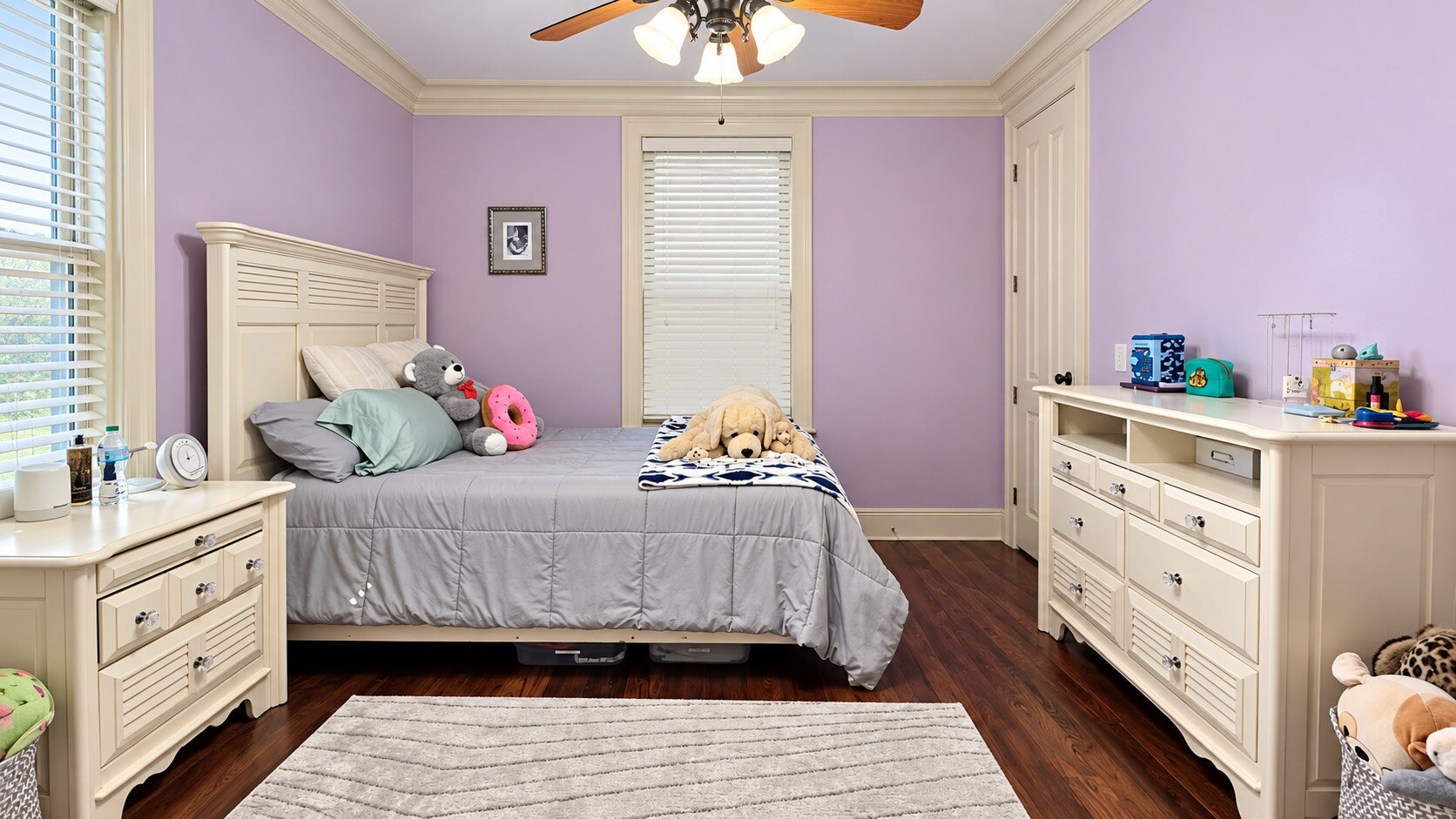Lavender bedroom with a neatly made bed, white dressers, and stuffed animals